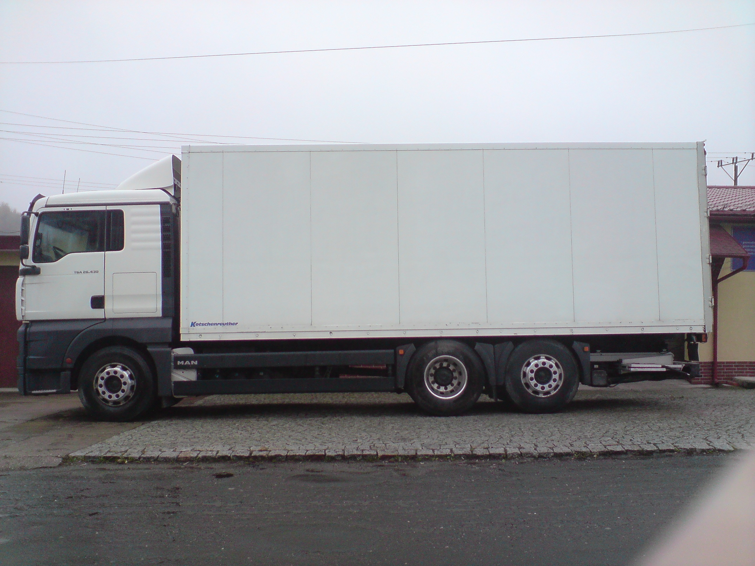 Biała ciężarówka MAN z zabudową kontenerową, widok z boku na tle pochmurnego nieba i fragmentu budynku z czerwoną dachówką.